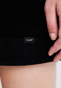 Svart kordtygsstoff med vertikala ribbor, som tydligt har en liten etikett med varumärket "Lee" nära fållen. Slät textur.