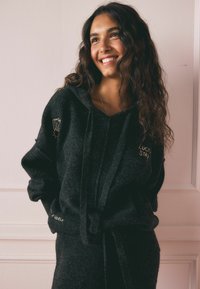 Schwarze Fleece-Hoodie mit lockerer Passform, ausgestattet mit goldenen Nähten an der linken Schulter und dem Ärmelbündchen. Weicher Stoff mit einer Kapuze zum Zuziehen.