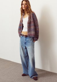 Cardigan tricotat multicolor cu închidere cu nasturi, maiou alb, blugi largi de culoare bleu deschis și pantofi slip-on maro pe un fundal neutru.