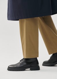 Fekete bőr csizmák vastag talppal, bézs egyenes szárú nadrággal viselve. A nadrág textúrált anyagú és letisztult kivitelű.