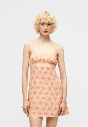 Ξανθιά γυναίκα που φορεί αμάνικο, ροδακινί φόρεμα με επαναλαμβανόμενο μοτίβο πορτοκαλί ηλιαχτίδας, στέκεται μπροστά σε ένα απλό φόντο.