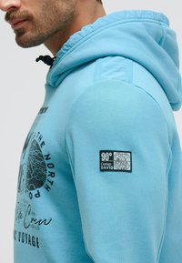 Light blue hoodie featuring a drawstring hood, front graphic print, and a black patch label on the left sleeve with a globe design.