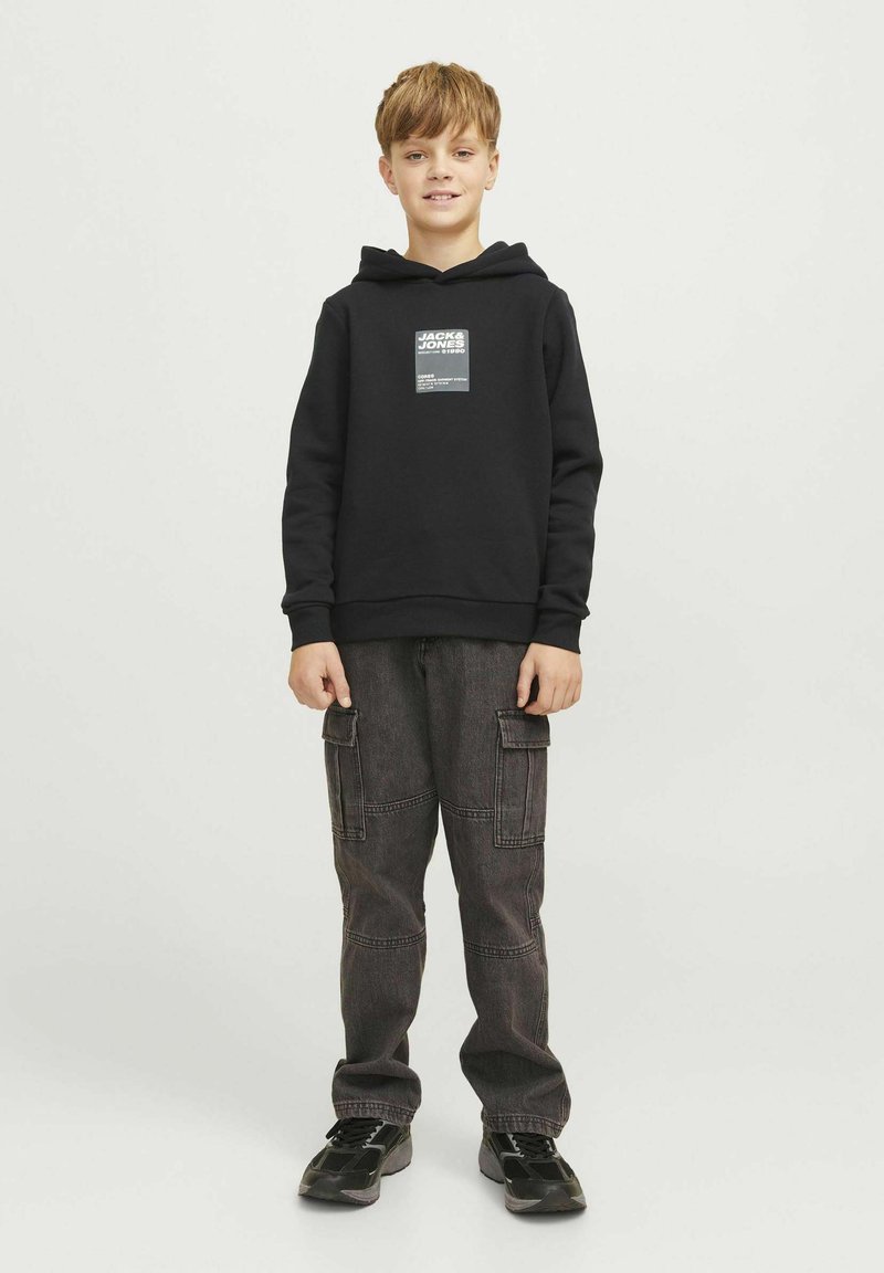 Felpa nera con cappuccio e patch logo frontale, abbinata a pantaloni cargo grigio scuro con molte tasche. Indossata con scarpe da ginnastica nere.