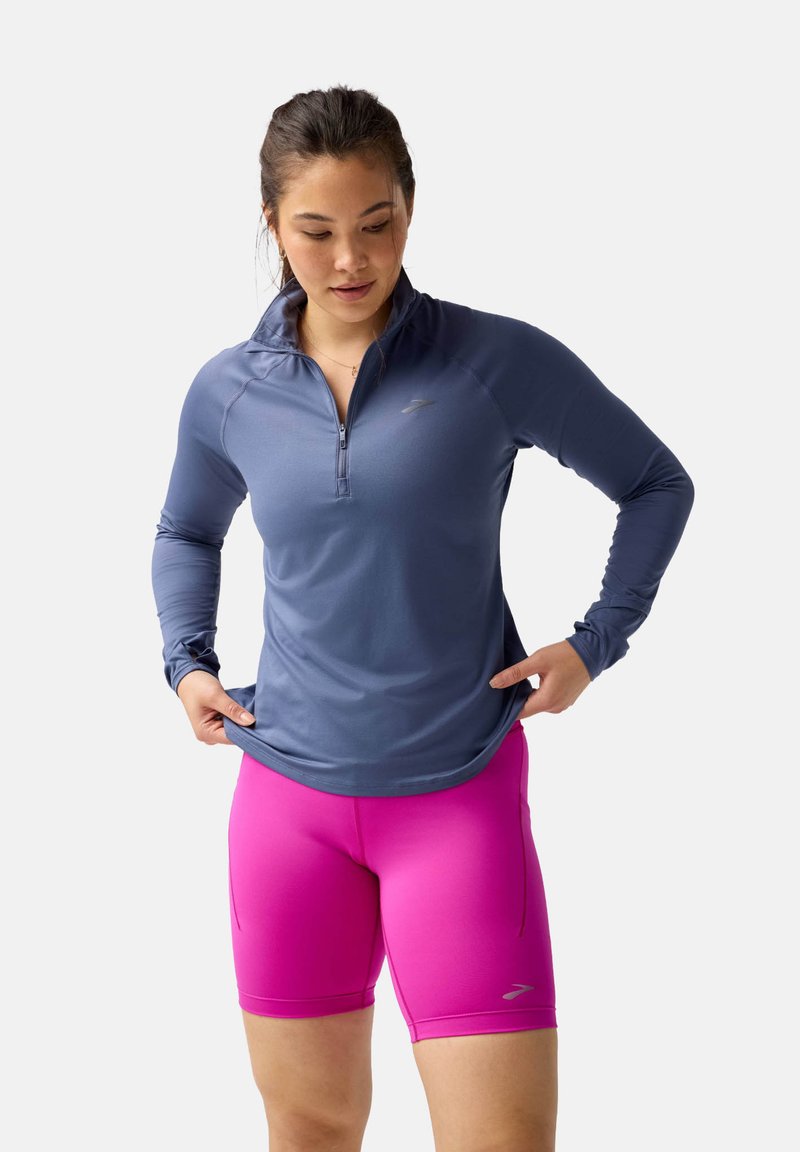 Maglia atletica a maniche lunghe con zip di colore blu, abbinata a pantaloni corti aderenti di colore rosa. Entrambi sono progettati per l'abbigliamento sportivo con materiale stretch.