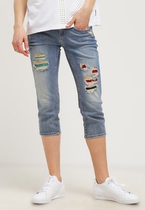 Personne portant un jean bleu délavé coupé avec des patchs en tissu colorés et des baskets blanches, debout avec une main dans la poche.