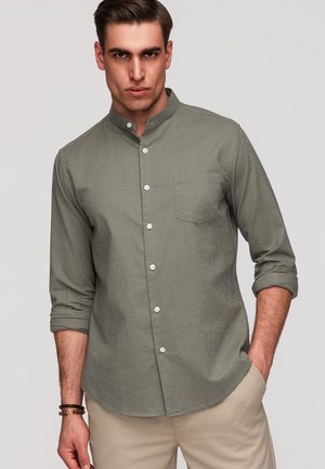 Mężczyzna w oliwkowej koszuli na guziki z długim rękawem i stojąc z jedną ręką w kieszeni, ubrany w beżowe spodnie.