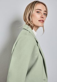 Blazer in lana verde chiaro con design strutturato, rever a punta e texture liscia, visto di lato su uno sfondo semplice.