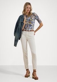 Top con stampa floreale in blu, beige e giallo; jeans bianchi aderenti; giacca di jeans drappeggiata sulla spalla; sandali con piatto marroni e cinturino alla caviglia.