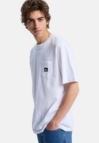 Hvid bomulds t-shirt med en afslappet pasform, der har en brystlomme og et lille sort logo patch. Påsyede ærmer og glat overflade.
