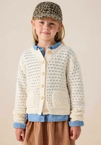 Weißer, gestrickter Cardigan mit einem strukturierten Muster, fünf goldenen Knöpfen und zwei Vordertaschen, getragen über einem blauen Jeanshemd und einem braunen Rock.