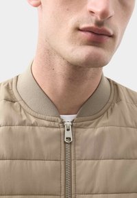 Chaqueta bomber beige acolchada con cuello de canalé, cierre de cremallera y textura suave, con costuras horizontales que añaden detalle.