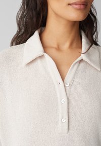 Ljusbeige stickad polo-shirt med krage, med en knapp-up framsidan och en texturerad tyg. Knapparna är vita och glansiga.