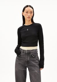 Schwarzes, kurzes Langarmoberteil mit gerippter Textur und kleinem Logo auf der Brust, kombiniert mit einem hellen Hemd und hochtaillierten grauen Jeans.