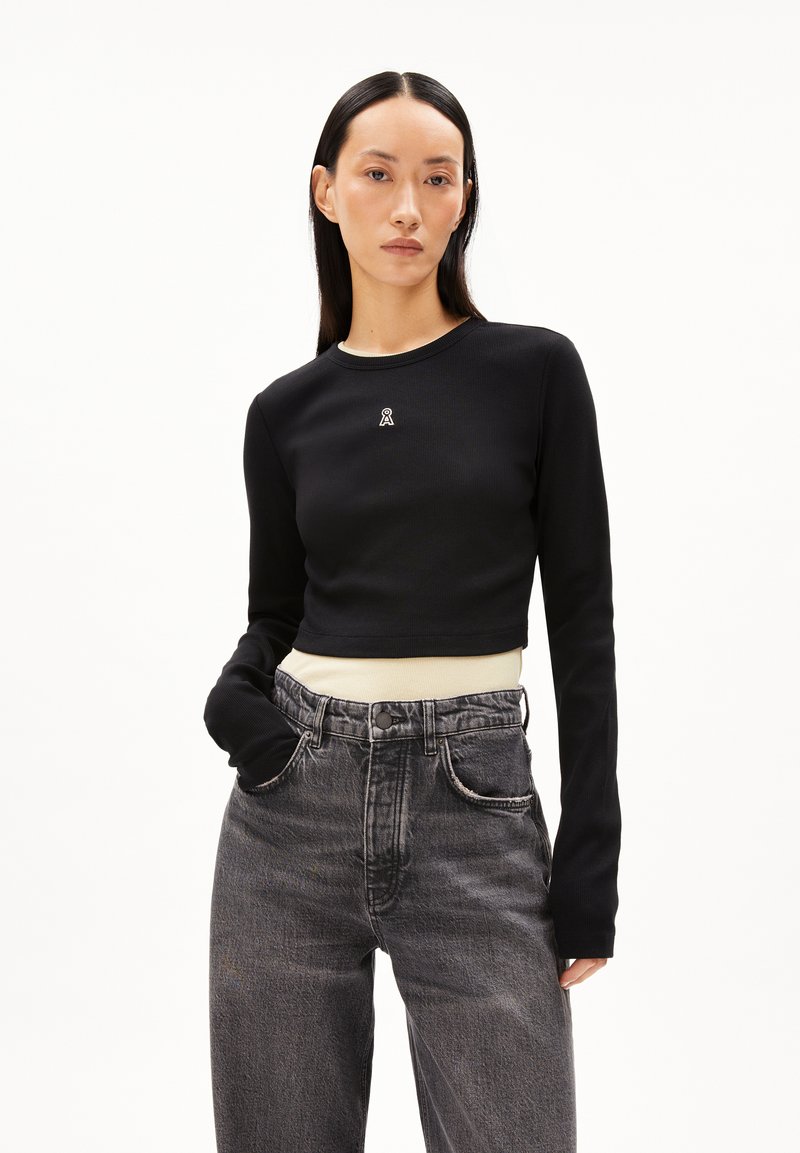 Schwarzes, kurzes Langarmoberteil mit gerippter Textur und kleinem Logo auf der Brust, kombiniert mit einem hellen Hemd und hochtaillierten grauen Jeans.