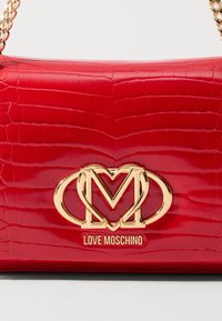 Borsa a mano in pelle verniciata rossa con texture coccodrillo, dotata di logo in tonalità oro e tracolla a catena. Forma rettangolare compatta.
