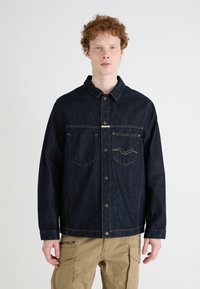 Mørkeblå denimjakke med knaplukning foran, to brystlommer og kontrasterende gule syninger. Har spids krave og afslappet pasform.