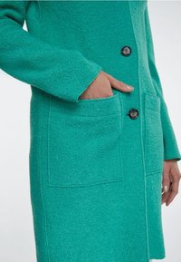Manteau texturé teal avec des boutons sur le devant et de grandes poches à patch ; poche intérieure à main gauche, main droite détendue sur le côté.