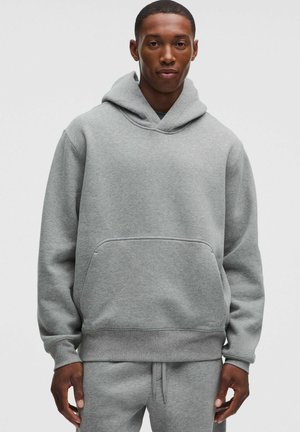 Junger Mann trägt einen schlichten grauen Hoodie und passende Jogginghose, steht vor einem weißen Hintergrund, schaut entspannt nach vorne.