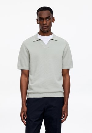 Homme portant un polo en maille à manches courtes gris clair par-dessus une chemise blanche et un pantalon foncé, debout devant un fond blanc uni.