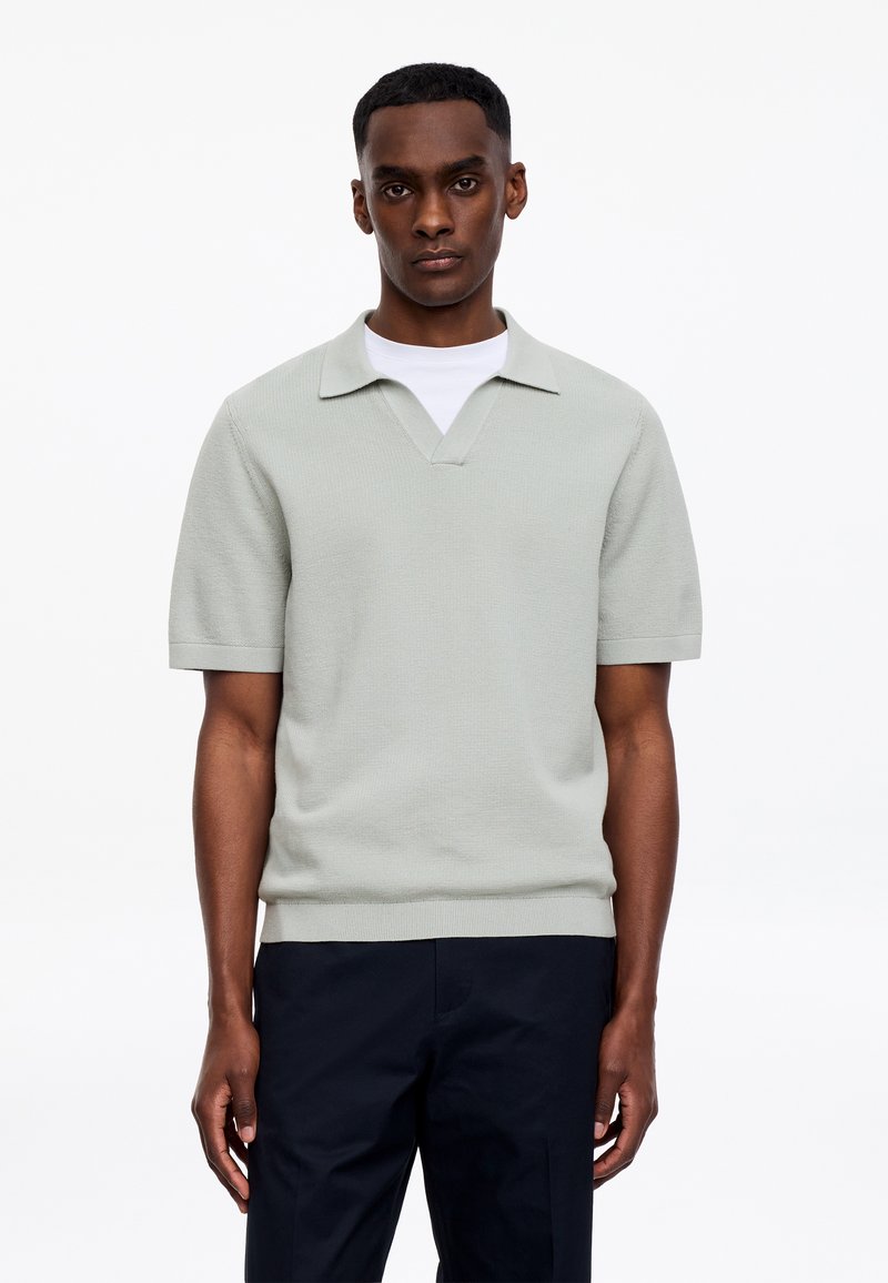 Homme portant un polo en maille à manches courtes gris clair par-dessus une chemise blanche et un pantalon foncé, debout devant un fond blanc uni.