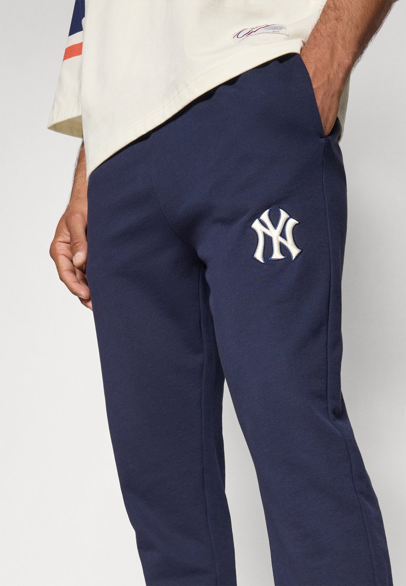 Man in marineblauwe trainingsbroek met wit New York Yankees-logo en beige T-shirt, staand met één hand in zijn broekzak.