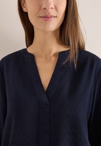 Marinebluse med tekstureret mønster, V-udskæring og korte ærmer. Stoffet har et blødt, glat udseende. Minimalistisk design.