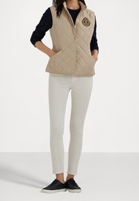 Beige prošívaná vesta s vysokým límcem, předními patentkami a emblémem na levé hrudi, spárovaná s bílými kalhotami a černými teniskami.