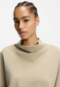 Beiges Rollkragenoberteil mit weicher, glatter Textur, dreieckigem Ausschnittdetail und subtilen Nähten, getragen von einem Modell mit Zöpfen.