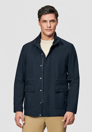 Mężczyzna w ciemnoniebieskiej kurtce zapinanej na zatrzaski z wieloma kieszeniami, na jasnobeżowym swetrze i beżowych spodniach, stojący na jednolitym tle.