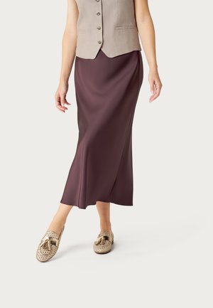 Mujer con un chaleco beige abotonado, una falda larga de color púrpura oscuro y mocasines beige tejidos con borlas, de pie contra un fondo liso.