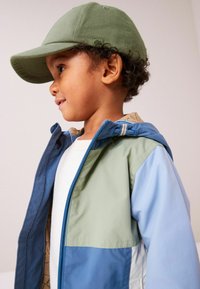 Cappello verde su un bambino con i capelli ricci. Il bambino indossa un giubbotto multicolore con pannelli blu e verdi e un interno a fantasia color beige.