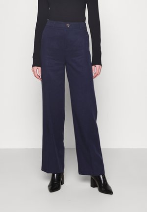 Vrouw draagt marineblauwe wijde broek, zwart top met lange mouwen en zwarte enkellaarsjes met hak, staand tegen een eenvoudige lichte achtergrond.