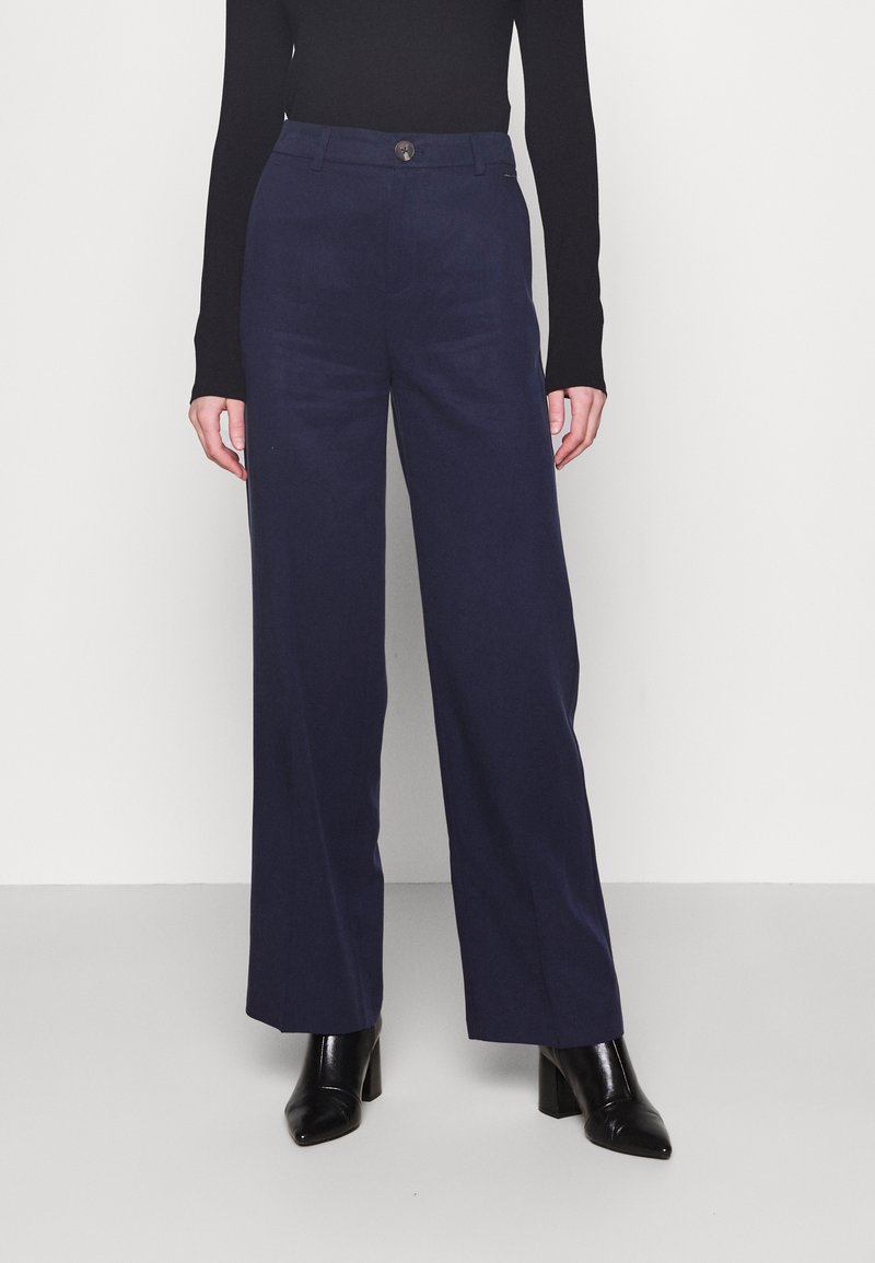 Femme portant un pantalon large marine, un haut noir à manches longues et des bottines à talons noires, debout devant un fond clair uni.