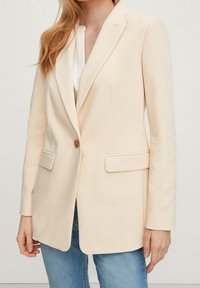 Blazer rosa chiaro con rever a intaglio, chiusura a un bottone, due tasche frontali e tessuto morbido, abbinato a un top bianco.