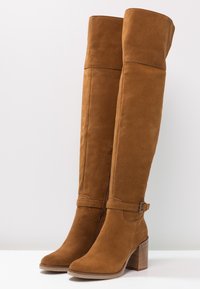 Botas de ante marrón por encima de la rodilla con tacón grueso, que presentan un acento de hebilla cerca de la parte superior y una textura suave.