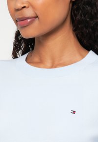 Gaiši zils apkakles džemperis, kas izgatavots no mīksta, gluda auduma, piedāvā nelielu logotipu ar sarkaniem un ziliem akcentiem uz krūtīm.