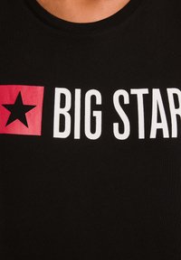 Tricou negru cu text alb „BIG STAR” și un pătrat roșu cu o stea neagră în partea stângă a pieptului.