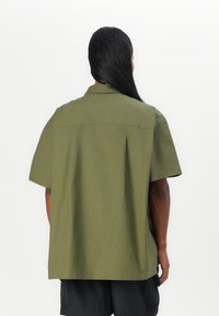 Short-sleeve, olive green shirt made of cotton. Features a relaxed fit, subtle seams, and a classic collar. Back view shows pleating detail.