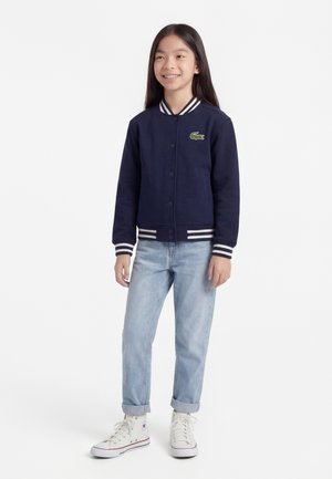 Młoda dziewczyna stojąca, ubrana w granatową kurtkę varsity z logo krokodyla, jasnoniebieskie dżinsy i białe sneakersy za kostkę.