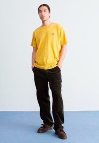 Gul bomulds T-shirt med korte ærmer og et logo på brystet, parret med mørkegrønne corduroy bukser og brune sko. Ensartet baggrund.