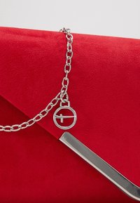 Pochette enveloppe en daim rouge avec bandoulière chaîne argentée et breloque logo circulaire argentée attachée près de la bordure métallique du rabat.