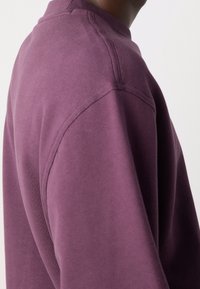 Fialová mikina s kulatým výstřihem, texturovanou látkou a hladkými švy. Boční pohled zdůrazňuje její volný střih a měkký materiál.