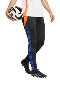 Schwarze Sporthose mit blauen Streifen und orangefarbenen Akzenten, kombiniert mit schwarzen Fußballschuhen. Der Träger hält einen schwarz-weißen Fußball.