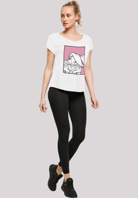 Hvit t-skjorte med korte ermer som har et tegneserie-design i en rosa ramme, kombinert med svarte leggings og svarte joggesko.