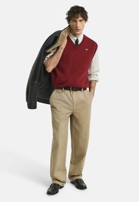 Roter V-Ausschnitt-Pullover über einem karierter Hemd, beige Hose und schwarze Lederschuhe. Das Model hält eine schwarze Jacke mit Lammfellfutter.