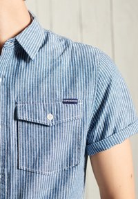 Short-sleeve blue and white striped shirt with a button-down collar, featuring a left chest pocket and a logo patch. Cotton material.