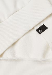 Tessuto bianco con texture a coste sul colletto, che mostra uno strato interno morbido. Un piccolo etichetta nera con un dettaglio grafico bianco è visibile.
