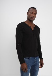 Zwarte langjarige henley-shirt met een V-hals en drie knopen, heeft een zachte textuur en blauwe accentdetails op de mouwen. G gedragen met blauwe jeans.
