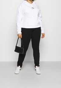 Felpa bianca con cappuccio, tasca frontale e logo, abbinata a pantaloni neri attillati e sneakers bianche. Piccola borsa nera tenuta in mano.