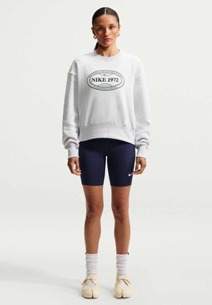 Kobieta ubrana w białą bluzę Nike 1972, granatowe krótkie spodenki rowerowe Nike, białe skarpetki i kremowe buty z rozdzielonym palcem, stojąca na jednolitym tle.